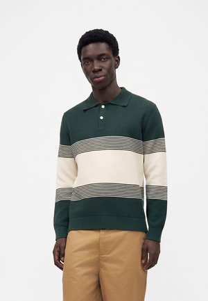 Jeune homme portant un pull polo à manches longues vert foncé et crème avec des rayures horizontales et un pantalon beige, debout devant un fond blanc uni.
