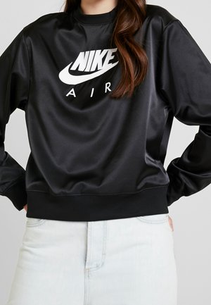 Osoba ubrana w czarną bluzę Nike Air z białym logo oraz jasnoniebieskie dżinsy, stoi z rękami na biodrach.