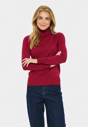 Burgunderfarbener Rollkragenpullover mit gerippter Textur und taillierter Passform. Zusammen mit blauen Jeans steht das Model vor einem hellen Hintergrund.