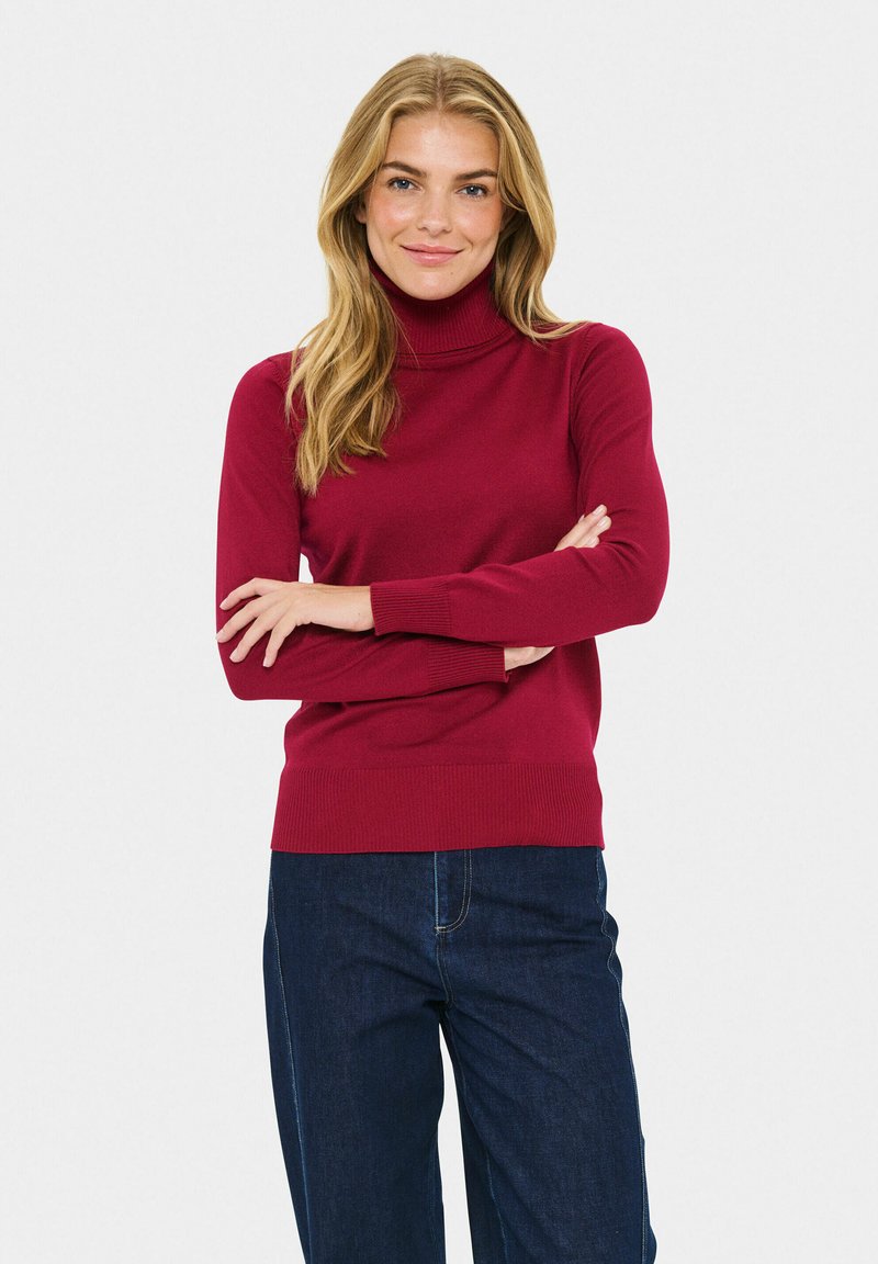 Burgunderfarbener Rollkragenpullover mit gerippter Textur und taillierter Passform. Zusammen mit blauen Jeans steht das Model vor einem hellen Hintergrund.