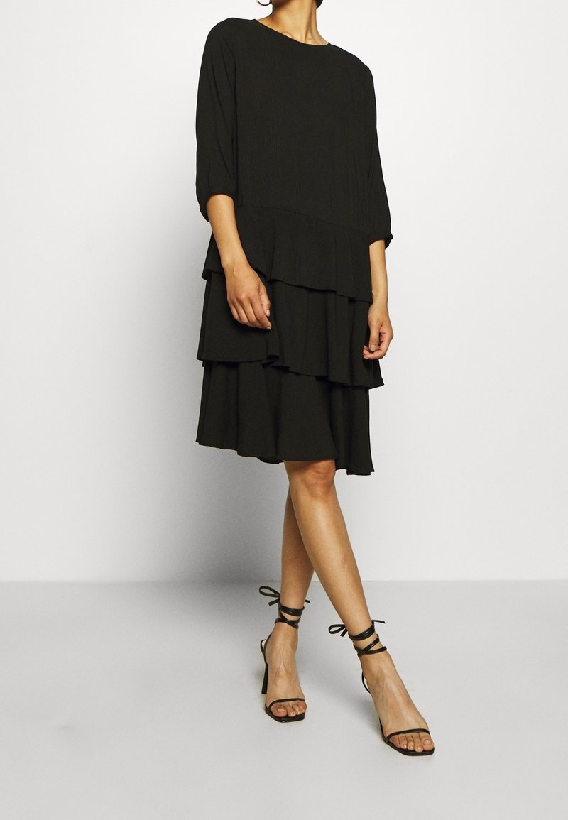Femme portant une robe noire à étages avec des manches aux coudes et des sandales à talons hauts noires attachées à la cheville, debout devant un fond uni.