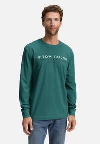 Uomo che indossa una camicia a maniche lunghe Tom Tailor di colore teal con testo bianco mentre è appoggiato a uno sfondo uniforme.