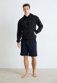Mann står barføtt iført svart Tommy Hilfiger hettegenser og marineblå shorts med merkevarer, mot en enkel hvit veggbakgrunn.