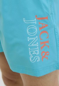 Bañadores turquesa en una tela ligera con un diseño gráfico vertical en blanco y coral de "JACK&JONES" en la pierna izquierda.