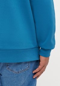 Blauwe sweatshirt met ribgebreide manchetten en zoom, met een soepele textuur. Draag het met spijkerbroeken, met een hand op de heup gelegd.