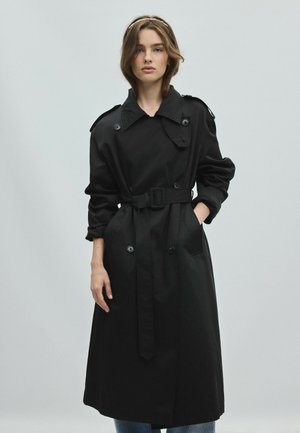 Jeune femme portant un trench-coat noir avec ceinture et manches retroussées, debout devant un fond gris clair uni.