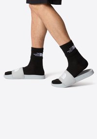 Chanclas grises con una banda ancha y acentos negros, usadas con calcetines negros que tienen un logo blanco. Material suave y liso y diseño plano.