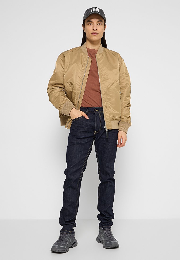 Veste bomber marron sur une chemise brune, associée à un jean bleu foncé et à des baskets grises. La personne porte une casquette sombre et se tient devant un fond neutre.