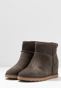 Par de botas de ante marrón oscuro hasta el tobillo con tacones de cuña y suelas de goma marrón claro, mostradas sobre un fondo claro y sin adornos.