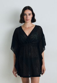 Abito in pizzo nero con profondo scollo a V, maniche corte e vita aderente. Il tessuto presenta un motivo intricato con ritagli ovali.