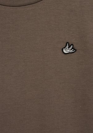 Hnedé bavlnené tričko s okrúhlym výstrihom a malým vyšívaným logom vtáka v bielej a čiernej farbe na ľavej strane.