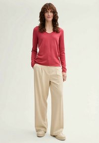 Pull à col en V à manches longues de couleur rose rouge, associé à un pantalon crème taille haute à jambes larges et des chaussures beiges, présenté sur un fond neutre.