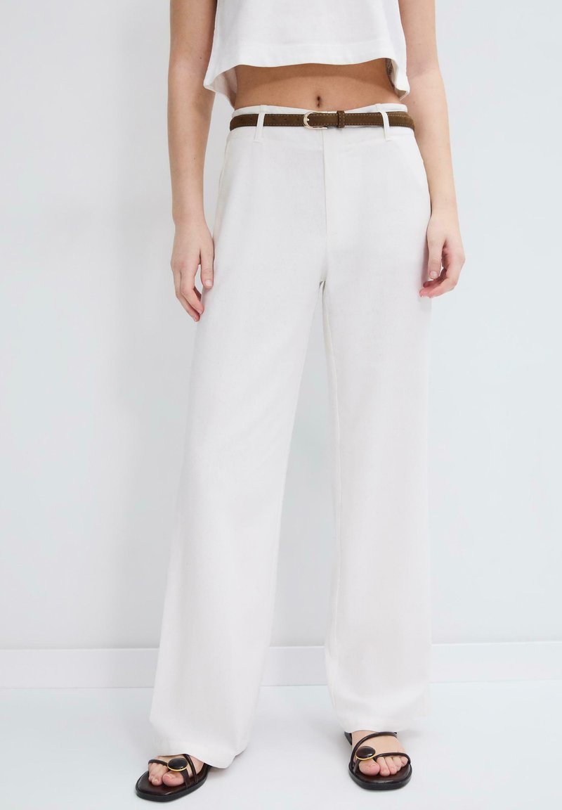 Donna con pantaloni bianchi a gamba larga e vita alta, cintura marrone e sandali neri aperti, in piedi su uno sfondo chiaro uniforme.