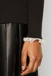 Svart ribbad långärmad topp med vit spetsruffel vid handleden, matchad med en svart plisserad imitationsläderkjol.