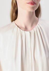 Blusa crema con scollo rotondo e pieghe, realizzata in un tessuto leggero e morbido. Nessun motivo visibile o dettagli hardware.