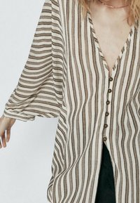 Bluzka w paski w kolorze beżowym i brązowym, z głębokim dekoltem w serek, z przodu na guziki i luźnymi rękawami. Materiał wydaje się lekki i ma miękką teksturę.