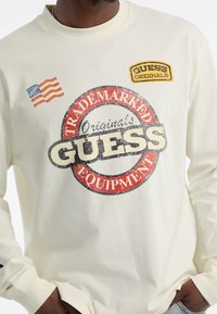 Langemouwige crèmekleurige shirt met een bedrukt rood en zwart rond logo, "GUESS Originals," en een blauwe Amerikaanse vlag detail op de linkerborst.