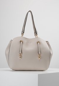 Vaaleanharmaa, teksturoitu nahkalaukku, jossa on kultaiset yksityiskohdat, kaksi olkahihnaa ja hienosti kaareva yläreuna, näytetään valkoisella jalustalla.