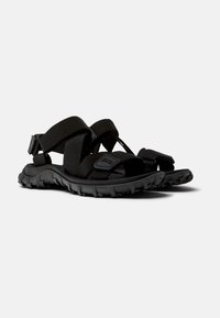 Sorte svarte sandaler med en tykk gummisåle, kross-remdesignet laget av vevd stoff, og justerbare borrelåsfester.