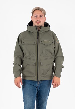 Jonge man in een olijfgroene hoodie met rits en flapzakken, gecombineerd met een blauwe spijkerbroek, staande tegen een witte achtergrond.
