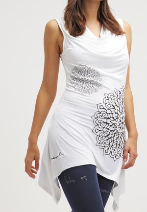 Mujer con túnica blanca sin mangas con diseños de mandalas negros, combinada con mallas azul oscuro con patrones de texto tenues.