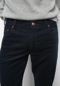 Marineblaue Cord-Hosen mit feinen vertikalen Streifen, ausgestattet mit einem Knopfverschluss, kupferfarbenen Beschlägen und vorderen Taschen, kombiniert mit einem grauen Pullover.