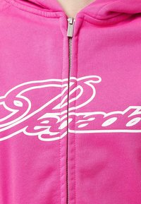 Sudadera con cremallera rosa hecha de tela suave, que presenta un diseño de logo blanco en la parte delantera. La cremallera de metal plateado se cierra verticalmente en el centro.