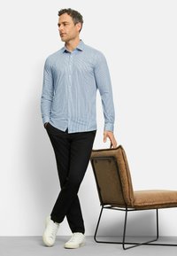 Hellblau gestreiftes Langarmhemd, schwarze Hose und weiße Sneaker, posiert neben einem braun gepolsterten Stuhl mit einem Metallrahmen.