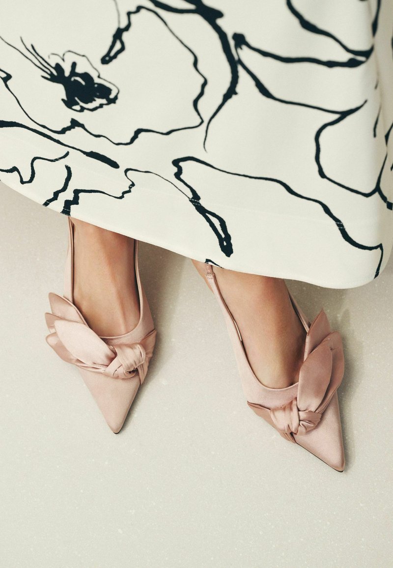Füße, die spitze, rosafarbene Satin-Ballerinas mit großen Schleifen tragen, teilweise bedeckt von einem weißen Rock mit schwarzem abstraktem Blumenmuster.