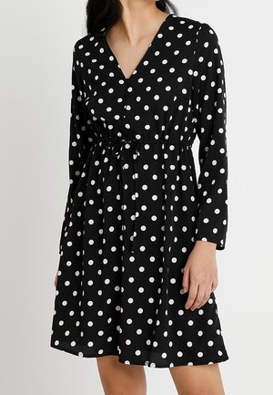 Kvinde iført en sort langærmet kjole med hvide prikker, V-hals og taljebånd, stående foran en ensfarvet baggrund.