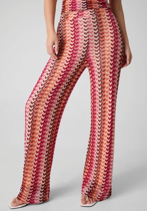 Primo piano di una persona che indossa pantaloni a vita alta e a zampa con un motivo a zigzag colorato lavorato a maglia in tonalità rosse, rosa, arancione e beige.
