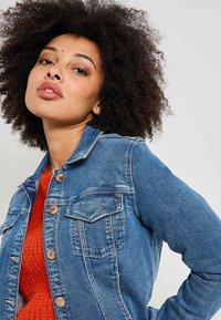 Chaqueta de denim en azul medio con un corte ajustado, que presenta dos bolsillos en el pecho y herrajes de botón de bronce. Tejido texturizado con un diseño clásico.