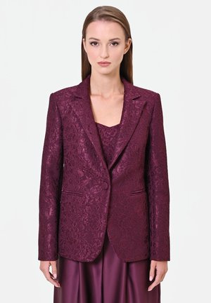 Donna con una giacca strutturata color viola scuro e una maglia abbinata, in piedi davanti a uno sfondo bianco uniforme, rivolta in avanti con un'espressione neutra.