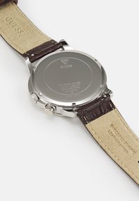 Bracelet en cuir marron avec texture embossée, fond de boîtier en acier inoxydable, résistant à l'eau et avec un logo clair. Design simple et fonctionnel.