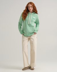 Giovane donna con lunghi capelli rossi che indossa un hoodie verde menta con loghi, pantaloni beige a gamba larga e sneaker marroni, in piedi davanti a uno sfondo chiaro.