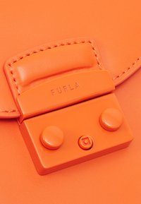 Zbliżenie zapięcia pomarańczowej skórzanej torby z wytłoczonym logo "FURLA" i dwoma okrągłymi guzikami z przodu.
