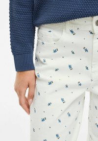 Une personne portant un pull en tricot bleu marine et un pantalon blanc à motifs floraux bleus, se tenant avec une position de main détendue.