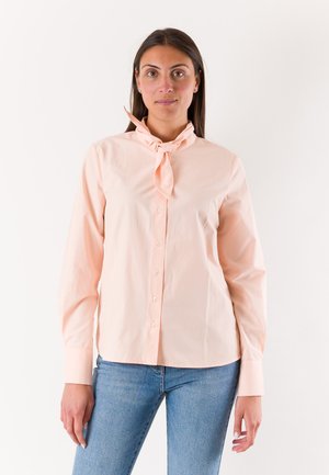 Donna con capelli scuri che indossa una camicetta rosa pallido con bottoni e dettaglio annodato al collo e jeans azzurri, in piedi davanti a uno sfondo bianco.