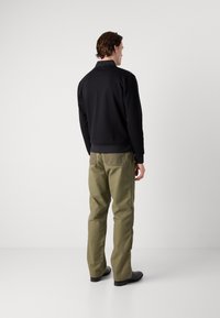 Veste zippée noire au design ajusté, associée à un pantalon vert olive. Texture lisse, détails minimaux et silhouette décontractée.