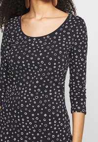 Černý top s dlouhým rukávem a kulatým výstřihem, na kterém se nachází vzor bílých kruhových tvarů různých velikostí po celé látce.