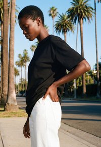 Mladić s kratkom kosom, obučen u crnu majicu i bijele hlače, stoji na pločniku uz cestu okruženu palmama u dnevnom svjetlu.