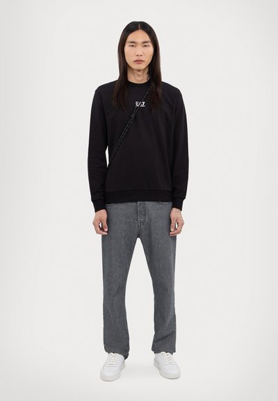 Sweatshirt noir à manches longues avec logo, pantalon en denim gris et baskets blanches. La tenue a une coupe décontractée et un design minimaliste.