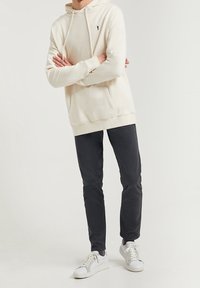 Krémová mikina s kapucí, přední kapsou a vyšívaným logem, doplněná tmavými kalhotami a bílými teniskami. Minimalistický a současný design.