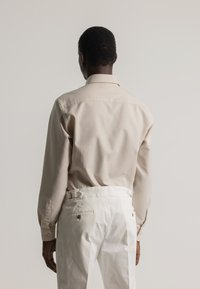 Hombre visto de espaldas con una camisa de manga larga beige metida en unos pantalones blancos de talle alto con bolsillos traseros y reguladores laterales.