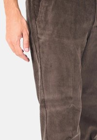 Pantaloni in corduroy marrone con striature verticali strette, in primo piano sulla trama del tessuto e una mano appoggiata sulla gamba.