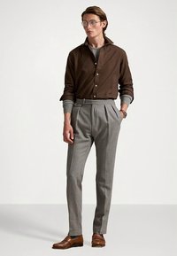 Brun skjorte med knapper, grå plissert bukse og brune skinnsko. Modellen har på seg runde briller og viser frem en klassisk, skreddersydd passform.