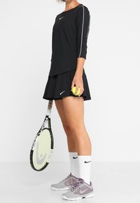 Schwarzes langärmliges Sportoberteil mit weißen Akzenten, schwarzer plissierter Rock, graue Turnschuhe, hält einen Tennisball und einen Schläger.