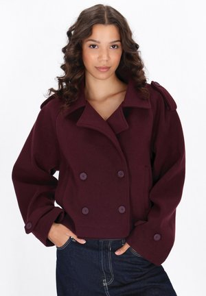 Manteau double-breasted bordeaux en tissu doux avec de gros boutons, grands revers et design court. Associé à un jean en denim foncé.