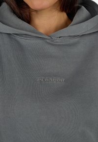 Γκρίζα φούτερ φτιαγμένο από μαλακό ύφασμα, διαθέτοντας τσέπη καγκουρό, κουκούλα με κορδόνι και ανάγλυφο λογότυπο "Pecador" στο στήθος.