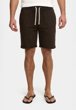 Mand iført mørkebrune shorts med snøring og sorte flip-flops, stående mod en ensfarvet baggrund.